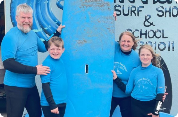 Family in blue surf school wetsuits smiling beside a large blue surfboard outside a surf shop.