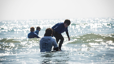 A surfing instructor in a white shirt helping a young male student practice standing up on a small surfboard in the sparkling ocean water.