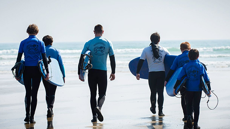 A group of six people, wearing wetsuits and carrying surfboards, walking away from the camera toward the ocean on a sunny beach.