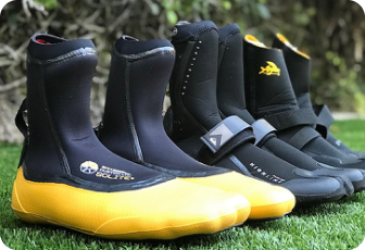 A row of several black neoprene wetsuit boots or booties, some with yellow toes and black straps, lined up on green grass outdoors.