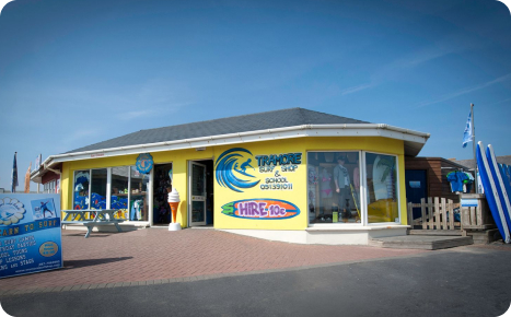 Interior view of a brightly lit surf shop featuring a wide array of merchandise including clothing, shoes, flip-flops, bags, and multiple surfboards hanging from the wooden ceiling and displayed on racks.