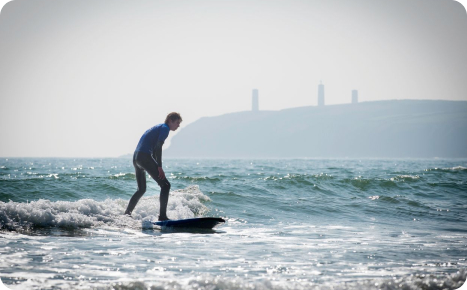 A male surfer in a blue wetsuit riding a dark surfboard on a small wave, with a foggy coastline and two tall structures visible in the distance.