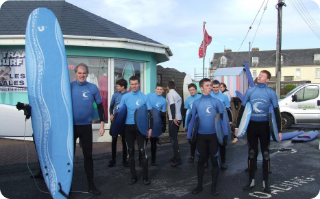 A group of nine male surfers and an instructor, all wearing blue surfing shirts and black wetsuits, standing with their blue surfboards outside the "Tramore Surf" shop on a street.