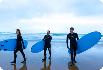 Three surfers in black wetsuits carrying bright blue surfboards, walking along a wet beach toward the ocean under an overcast sky.