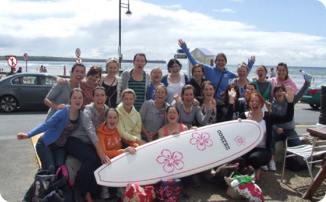 A large group of smiling women posing outdoors with a white surfboard decorated with pink flowers, with a beach visible in the background.