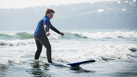 Beginner surfer in a wetsuit riding a small wave near the shore during a one-day surf camp.