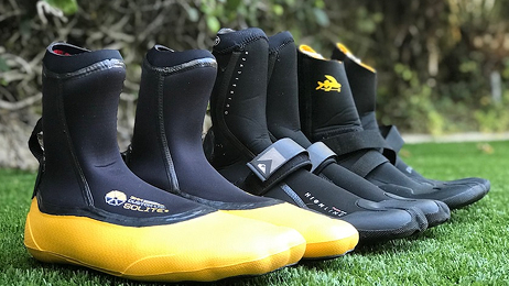 Water sports boots lined up on grass, including yellow-soled and black surf booties.