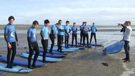College group in wetsuits standing on surfboards on the sand while an instructor gives directions.