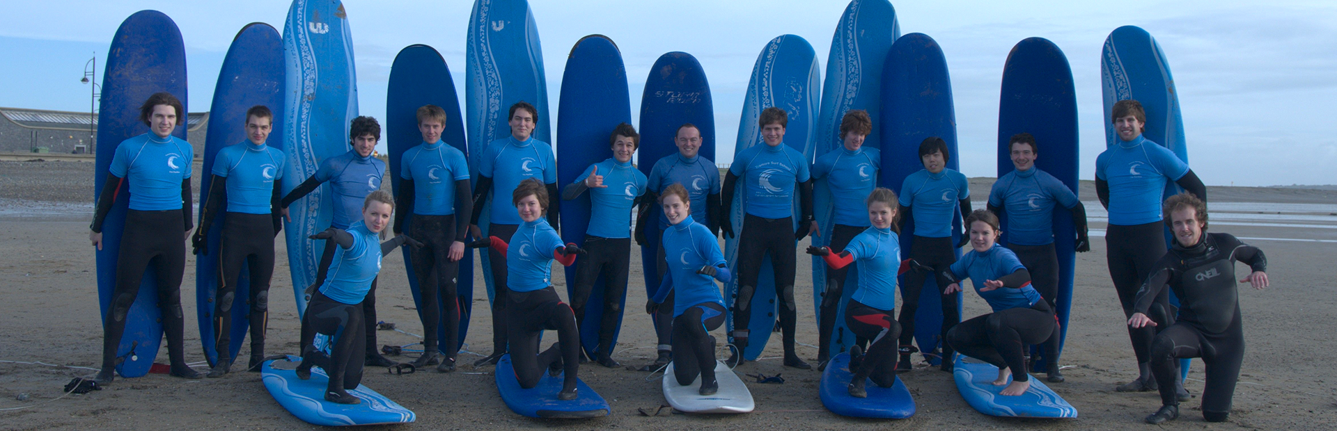 Large group of surfers in wetsuits posing on the beach holding surfboards during a group lesson.