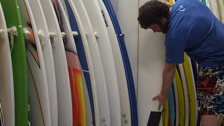 Man selecting a surfboard from a rack of upright boards inside a surf shop.