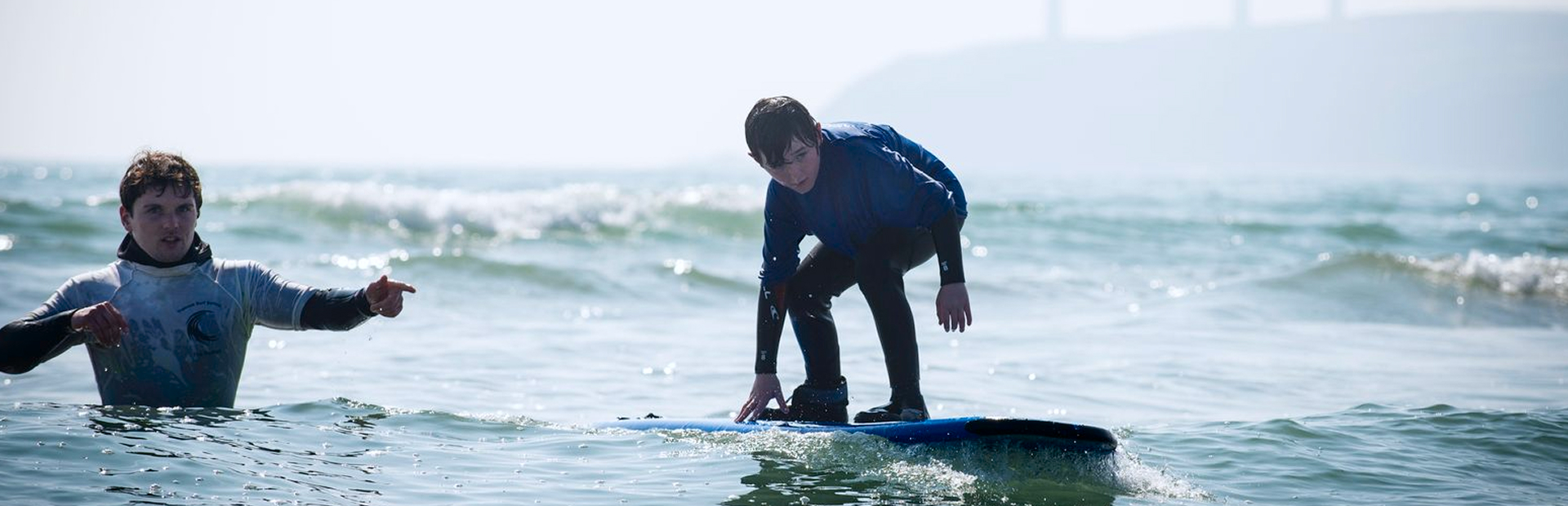 Child surfing a small wave while an instructor guides nearby in the water during a kids’ surf camp.