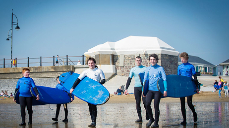 Group of beginner surfers in wetsuits carrying boards while walking along the beach during a surf lesson.