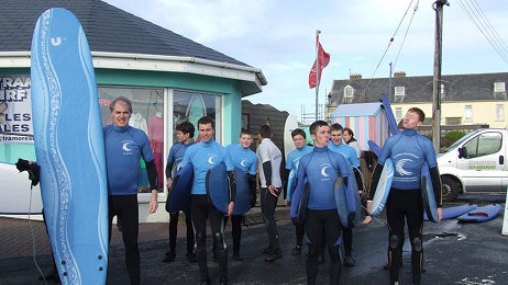 School group in wetsuits walking outside a surf shop carrying boards during a surf tour.