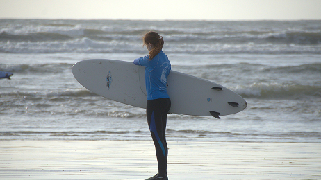 Surfer in a wetsuit carrying a surfboard while standing on the beach facing the ocean.