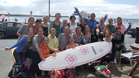 Group celebrating a hen party on the beach, posing with a surfboard and cheering.