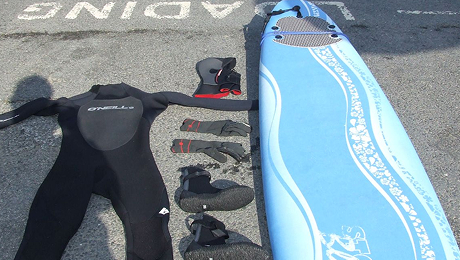 Surf hire package laid out on pavement, including a surfboard, wetsuit, gloves, boots, and hood.