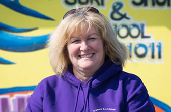 Portrait of the surf school owner in front of the shop wearing a purple hoodie in front of the shop sign.