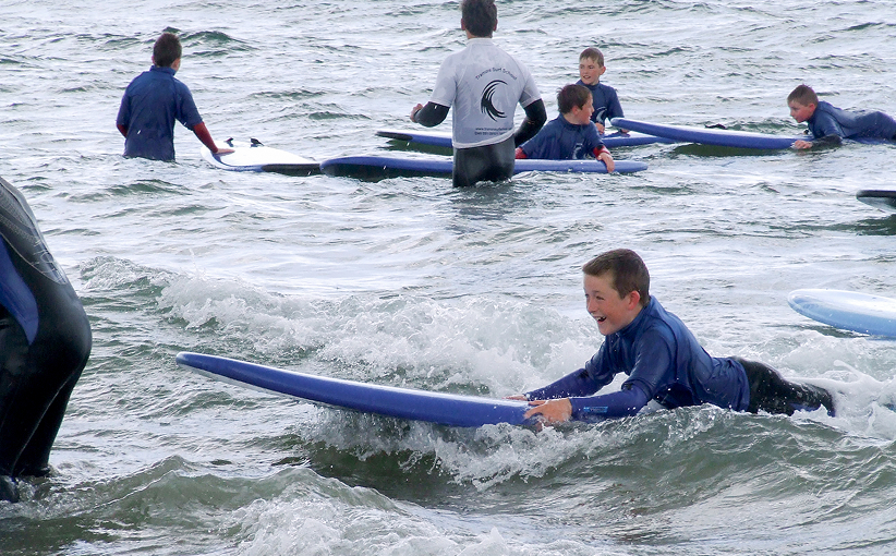 Children in wetsuits paddling on surfboards during a supervised surf lesson, smiling as small waves roll in.