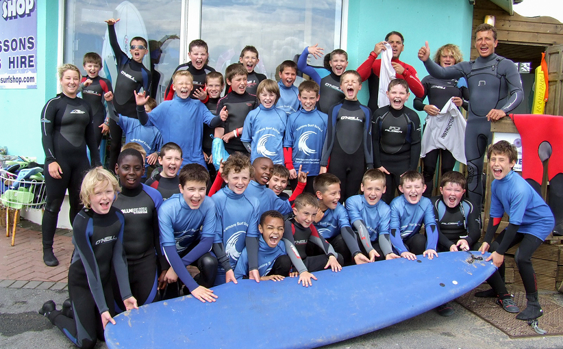 A large group of children in wetsuits and surf tops cheering and posing together outside a surf shop, gathered around a surfboard with instructors standing behind them.