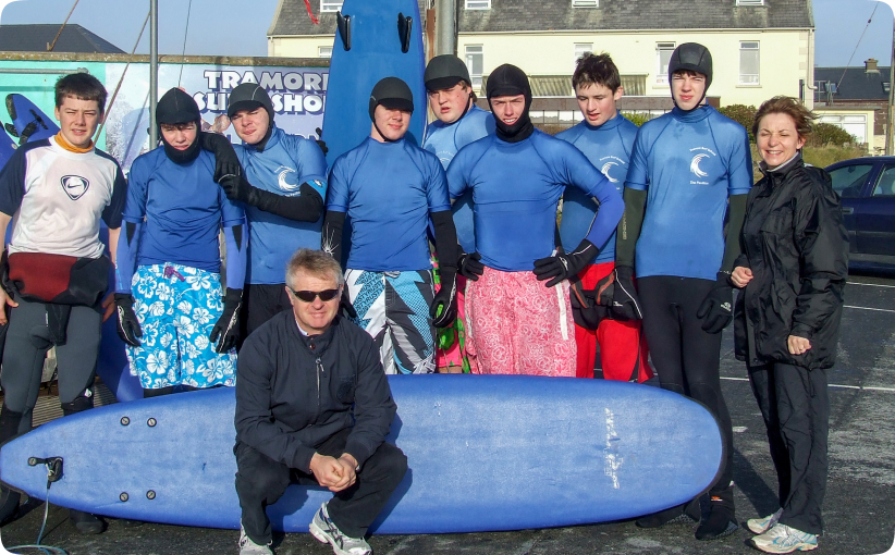 A group of teenagers in wetsuits and blue surf tops posing together with a large surfboard outside a surf school, accompanied by two adults, likely instructors or organisers.