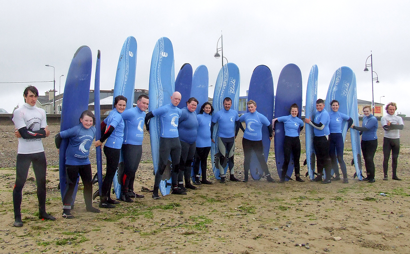 Group of surfers lined up in blue wetsuits posing on the beach with upright surfboards.