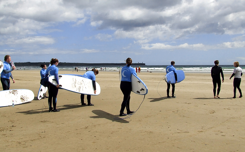 Group of surfers in wetsuits carrying surfboards across a sandy beach toward the sea.