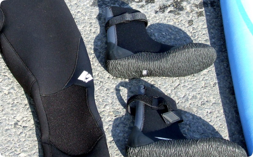 Neoprene surf boots and wetsuit leg placed on the ground beside a surfboard.