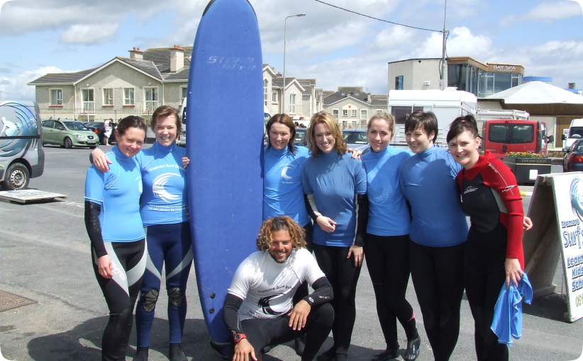 A college group in blue surf tops gathers around their instructor and a large surfboard outside the surf school, smiling after their lesson near the seafront.