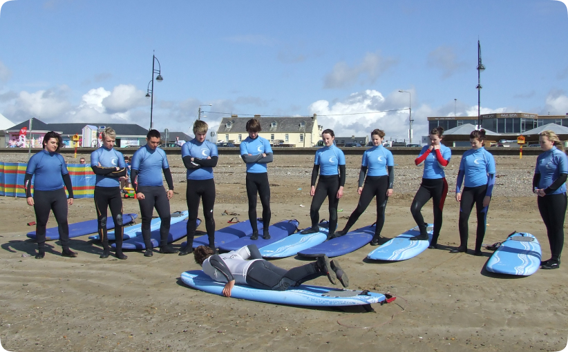 Surf instructor lying on a surfboard demonstrating technique to enter the sea while a group of students in blue wetsuits watch on the beach.
