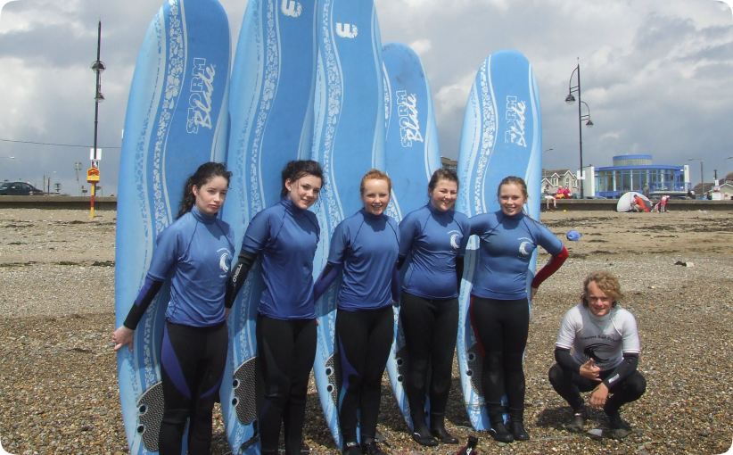 Five students in blue wetsuits standing in front of upright blue surfboards on a beach, with an instructor crouching beside them.