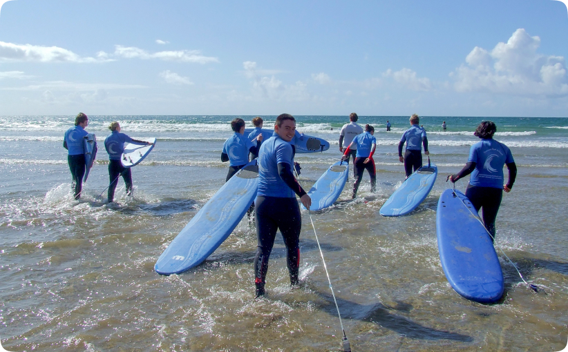 Group of surf students in blue wetsuits carrying surfboards into the sea during a lesson.
