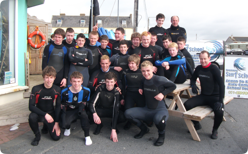 A college group in wetsuits poses together outside the surf school, smiling after their session and gathered around a picnic bench near the beachfront.