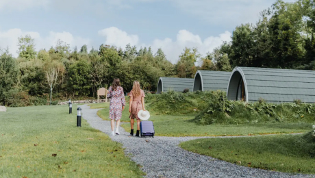 Two women walking along a gravel path toward Comeragh pod cabins surrounded by greenery