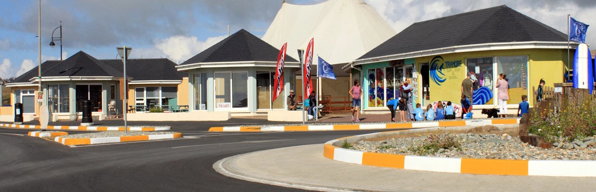 Seafront buildings including Tramore surf shop with people gathered outside near a roundabout and promenade area.