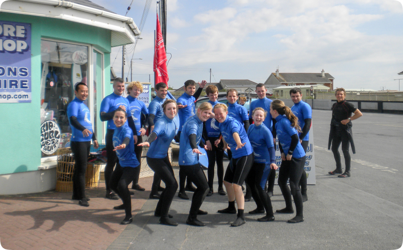 A cheerful corporate group in matching blue surf school tops and wetsuits gathers outside the surf shop, posing playfully before heading out for their surf lesson together.