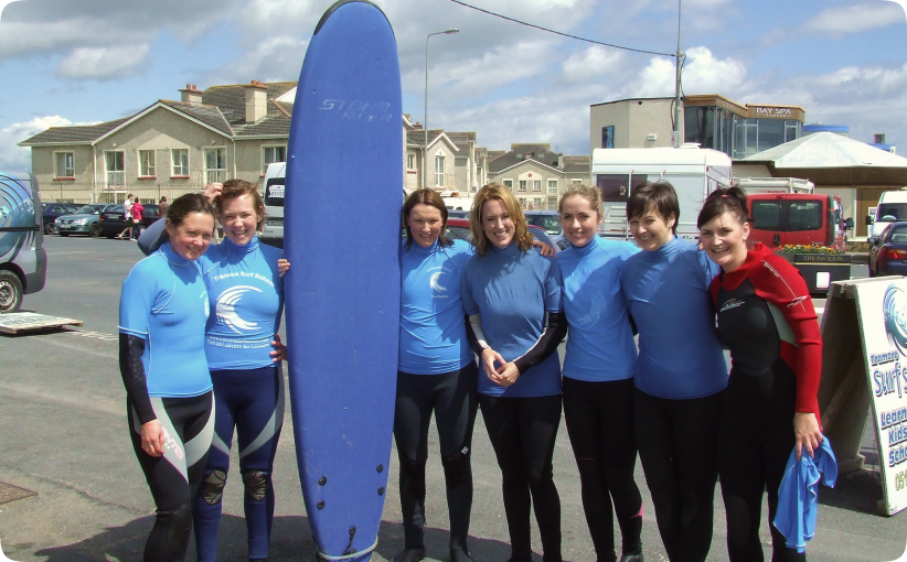 A corporate group of women in wetsuits and blue surf school tops standing together after their lesson, smiling beside a large surfboard in the car park near the beach.