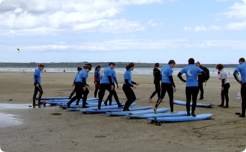 A corporate group in blue surf school tops and wetsuits practising balance and stance on surfboards laid out on the sand during a beach training session before entering the water.