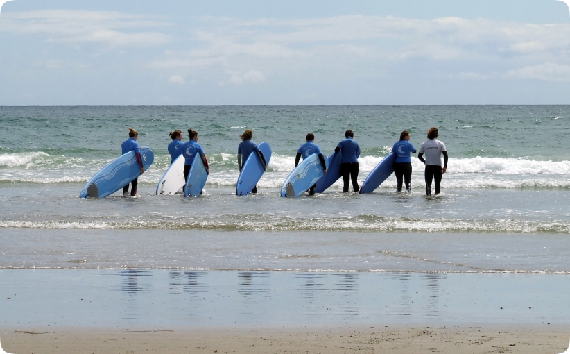 A corporate group in matching surf school tops carrying blue surfboards as they wade into the sea together, beginning their surf lesson under instructor supervision on a calm day.