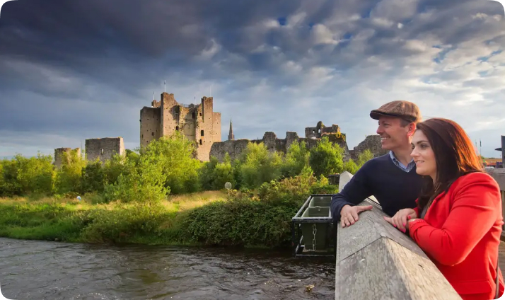 Couple standing on a bridge looking toward historic castle ruins beside a river under a dramatic sky.