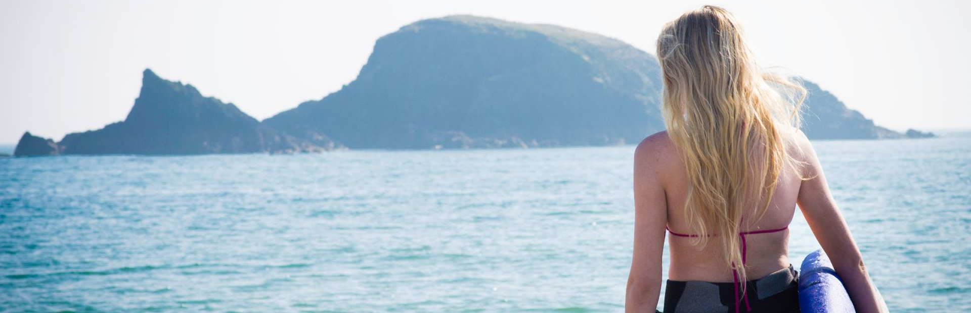 Woman with long blonde hair holding a surfboard and looking out at the sea toward a rocky island coastline.