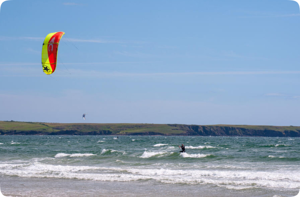 Kite surfer riding small waves near the shore with a colourful kite flying overhead and cliffs in the background.