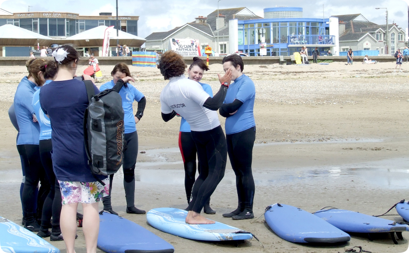 Surf instructor demonstrating technique to a group of adult learners standing beside boards on the beach.