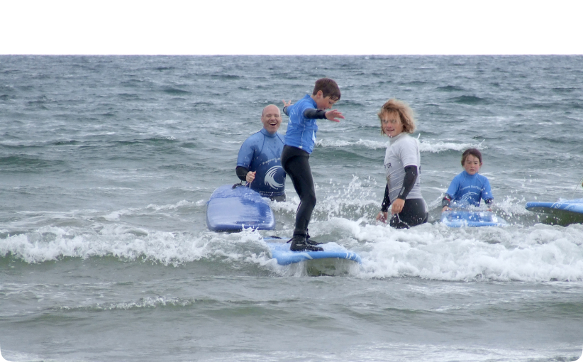 Child riding a small wave on a surfboard with instructors nearby during a family lesson.