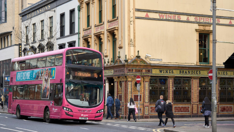 Pink double-decker Belfast bus driving past a corner pub on a busy Belfast street with pedestrians crossing.