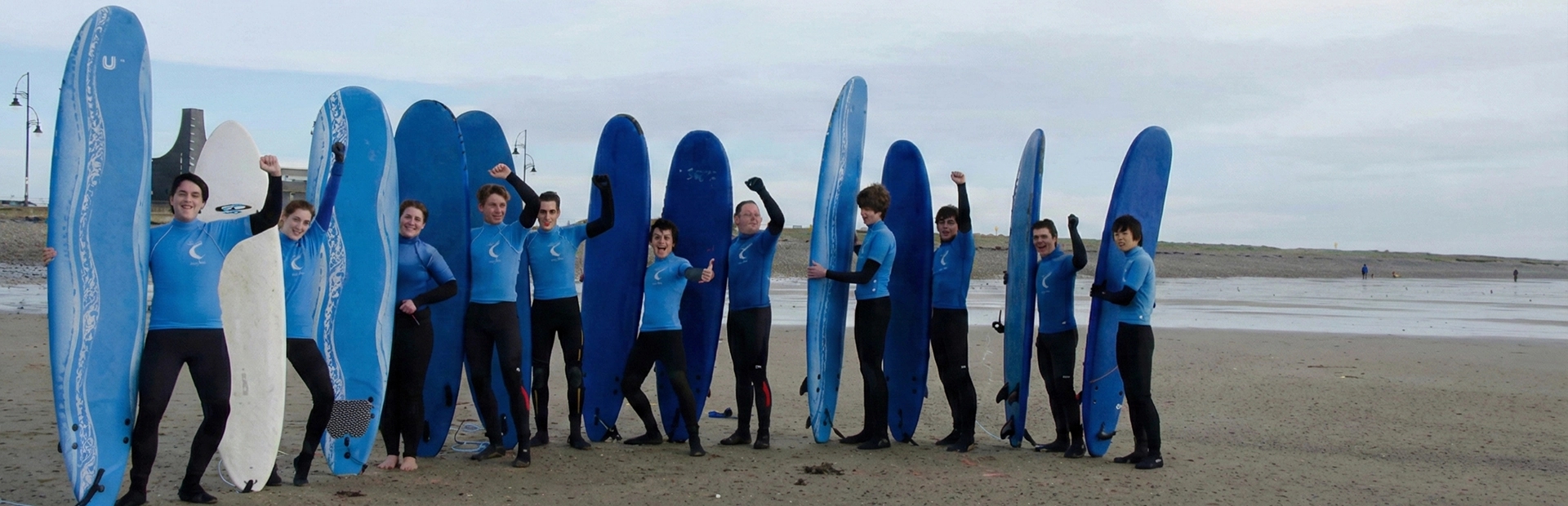 Group of surfers in wetsuits posing on the beach with upright surfboards.