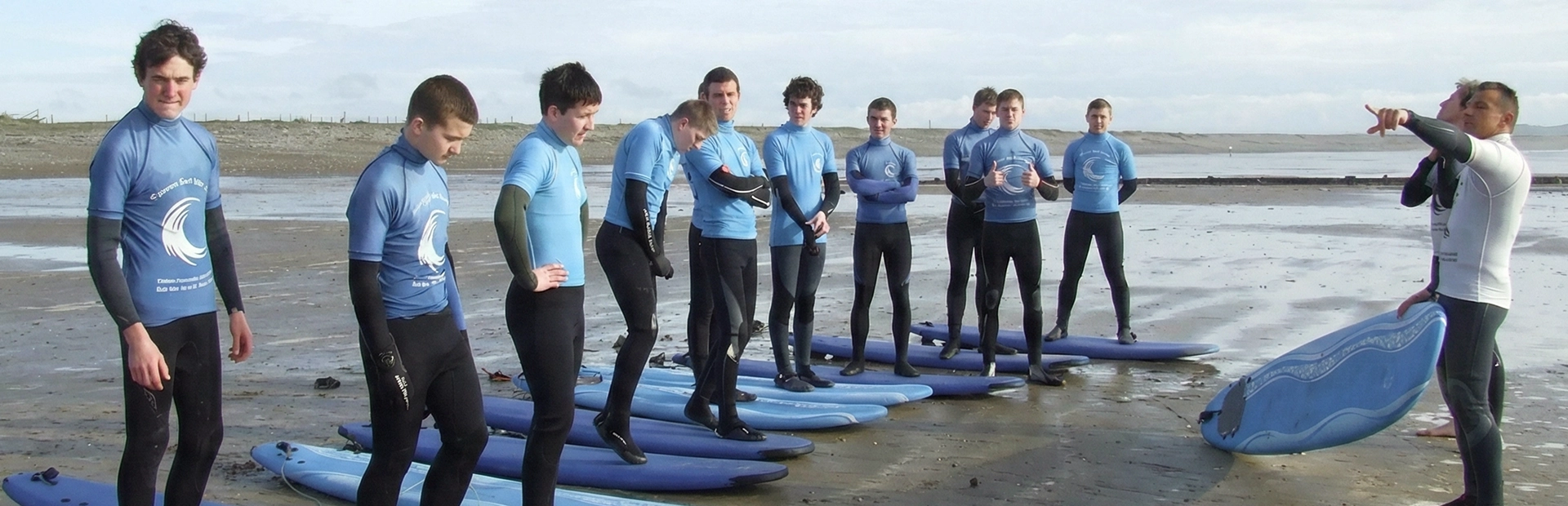 A group of participants in matching blue rash vests stands on their surfboards on the sand while an instructor demonstrates technique and gives directions, preparing them for entering the water.