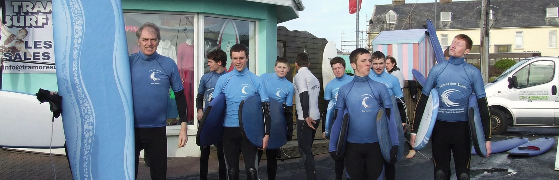 Group of surf students in blue wetsuits standing outside a surf shop holding surfboards before a lesson.
