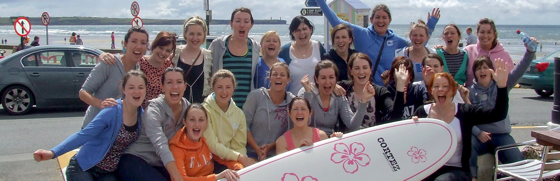 Group celebrating a hen party on the beach, posing with a surfboard and cheering.