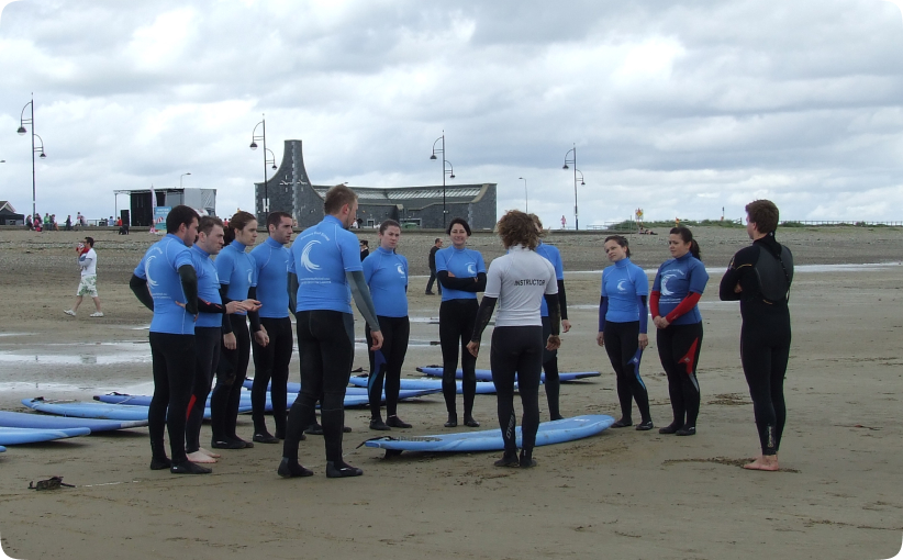 Surf instructor addressing a group of adult learners gathered around boards on the beach.