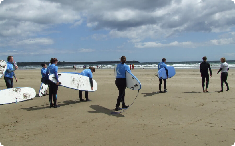 Group of surfers carrying boards across the beach toward the water during a lesson.